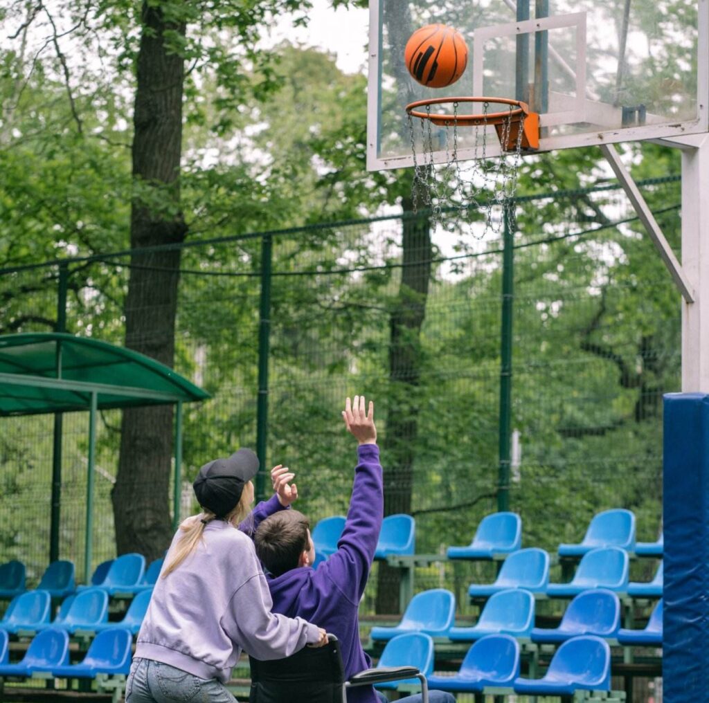 A Person With A Disability, Wearing A Purple Hoodie, Shoots A Basketball Toward A Hoop While Being Assisted By An Ndis Provider In A Baseball Cap And Gray Hoodie. They Are On An Outdoor Court With Blue Bleachers, Surrounded By Trees.