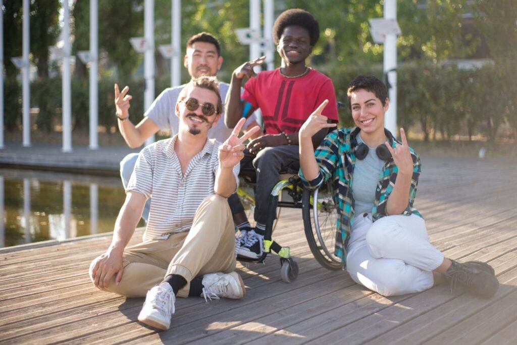 A Diverse Group Of Four Friends, Including A Person In A Wheelchair, Sit And Smile On A Wooden Dock Beside A Body Of Water. They Are All Making Peace Signs With Their Hands. The Background Features Greenery And A Sunny, Outdoor Setting.