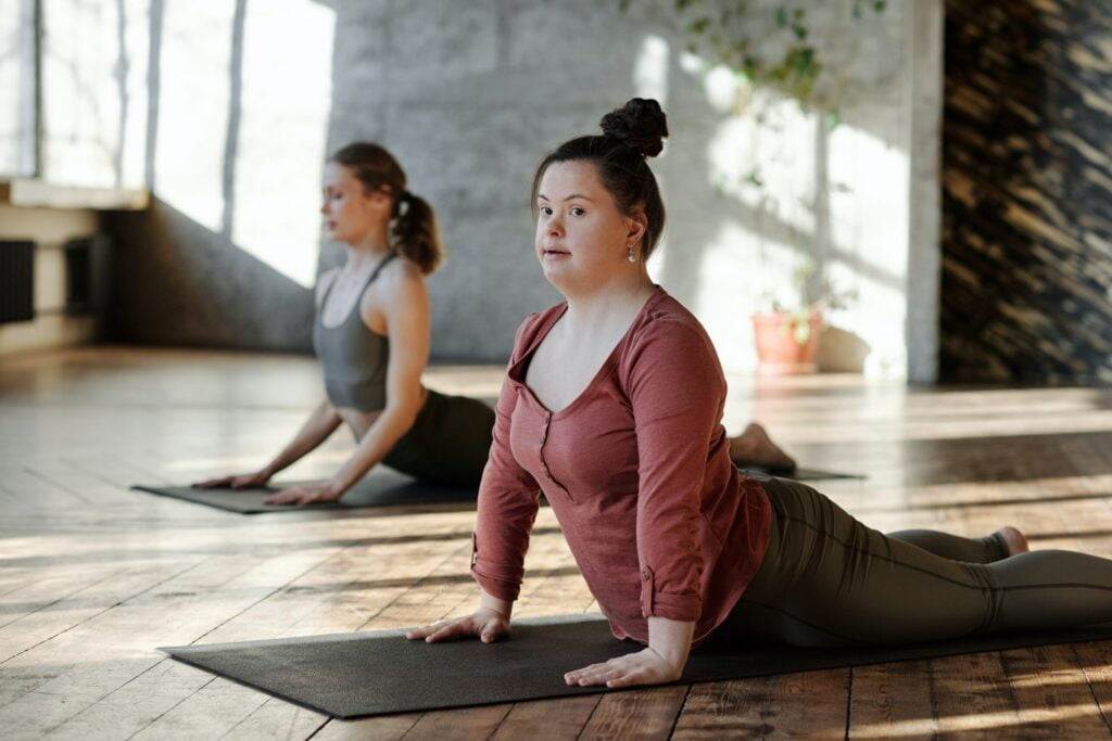 Two Women Practice Yoga Indoors On Mats In A Sunlit Room. The Woman In The Foreground Is In Cobra Pose, Wearing A Long-Sleeved Top And Leggings. The Woman Behind Her Is Also In Cobra Pose, Dressed In A Tank Top And Leggings. Plants And Light Stream Through The Windows.