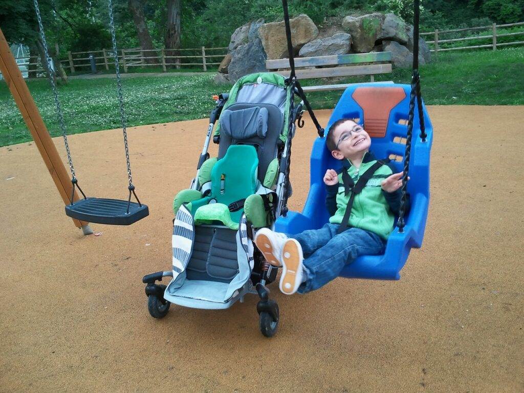 A Child Wearing Glasses And A Green Jacket Is Joyfully Swinging In A Blue Adaptive Swing Next To An Empty Regular Swing. A Stroller With A Green Seat Is Positioned Nearby On The Playground With A Grassy Area And Rocks In The Background.