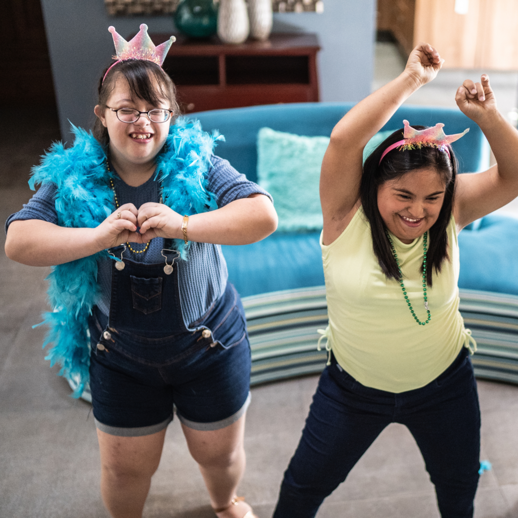 Two Young Women Are Joyfully Dancing In A Living Room During A School Holidays Program. One Wears A Blue Feather Boa, Overalls, And A Pink Crown With Her Hands Forming A Heart Shape. The Other Wears A Yellow Top, Jeans, And Similar Crown With Her Arms Raised In The Air. They Both Smile Excitedly.