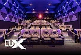 A Modern Movie Theater Interior Featuring Luxurious Reclining Seats Arranged In Rows On Multiple Levels. The Walls Have A Geometric Design, And The Lighting Is A Mix Of Soft Purple And Blue Hues. In The Bottom Left Corner, There Is A Logo That Reads &Quot;Movie Lux.