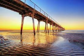 A Picturesque View Of A Wooden Pier Extending Into The Ocean During Sunset. The Sky Is Painted With Vibrant Shades Of Orange, Yellow, And Blue, While The Calm Water Beneath And Rippled Sand Reflect The Colorful Sky.