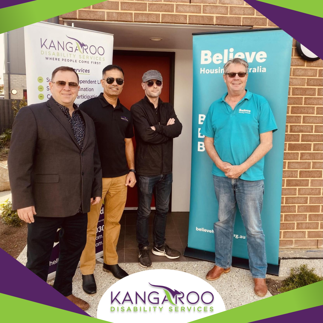 Four Men Stand Together Outside A Building, Smiling At The Camera. Two Banners, Prominently Featuring &Quot;Kangaroo Disability Services&Quot; And &Quot;Believe Housing Australia&Quot; Logos, Can Be Seen Behind Them. The Men Wear Different Attire, Ranging From Casual To Business Casual.