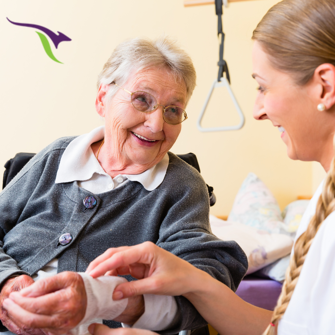 A Smiling Elderly Woman With Grey Hair And Glasses Sits In A Wheelchair, Holding The Hand Of A Young Female Caregiver With Blonde Hair. The Caregiver Is Also Smiling As She Tends To The Woman'S Hand, Representing The High Intensity Clinical Supports They Provide. There Is A Triangular Logo On The Top Left Corner.