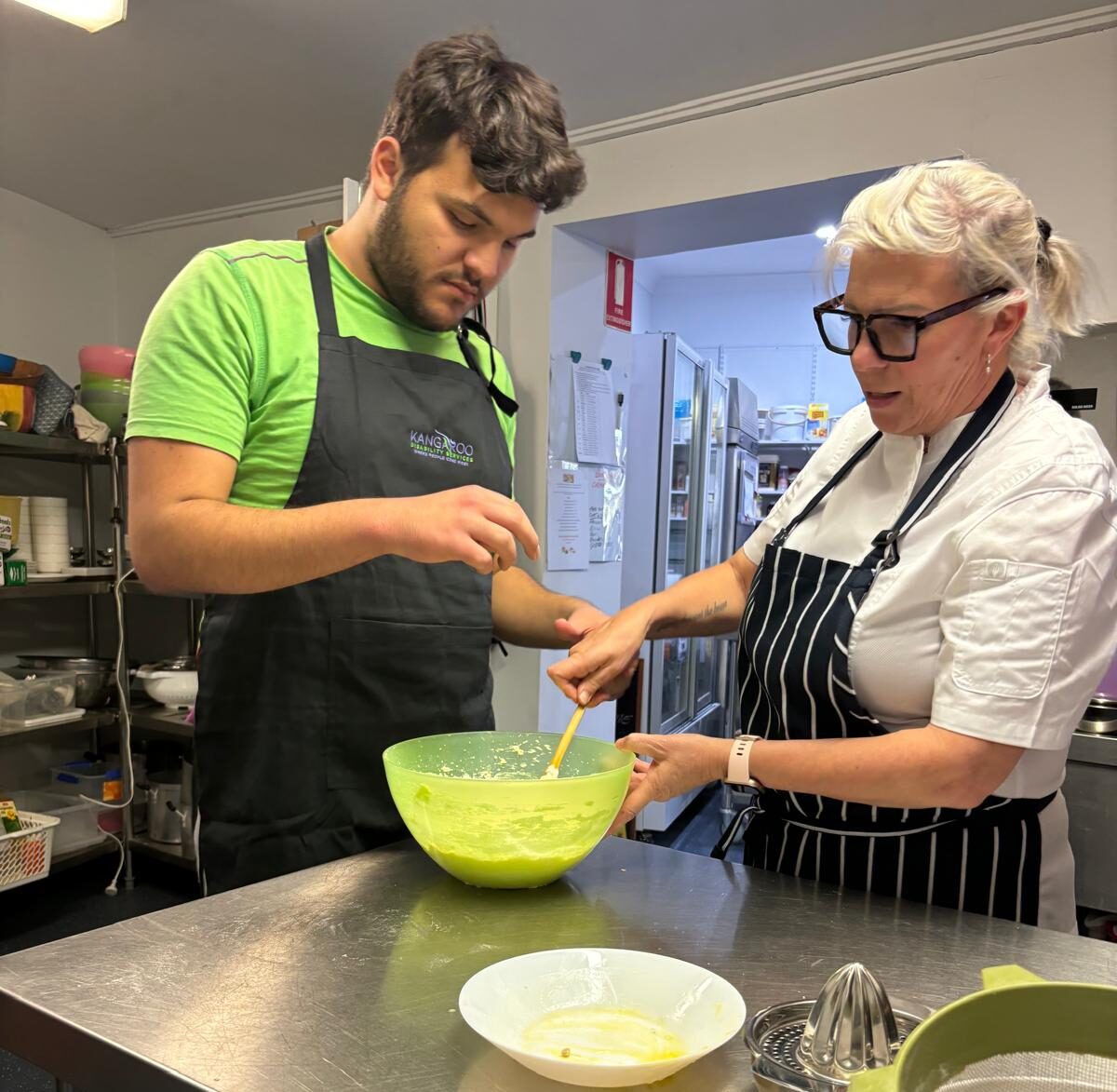 Two People In A Kitchen, One Wearing A Green Shirt And Black Apron Stirring Contents In A Green Bowl, While The Other, Wearing Glasses And A Black Striped Apron, Assists. A Juicer, Bowl, And Other Kitchen Tools Are On The Stainless Steel Counter During Their Week 2 Cooking Classes At Kds.