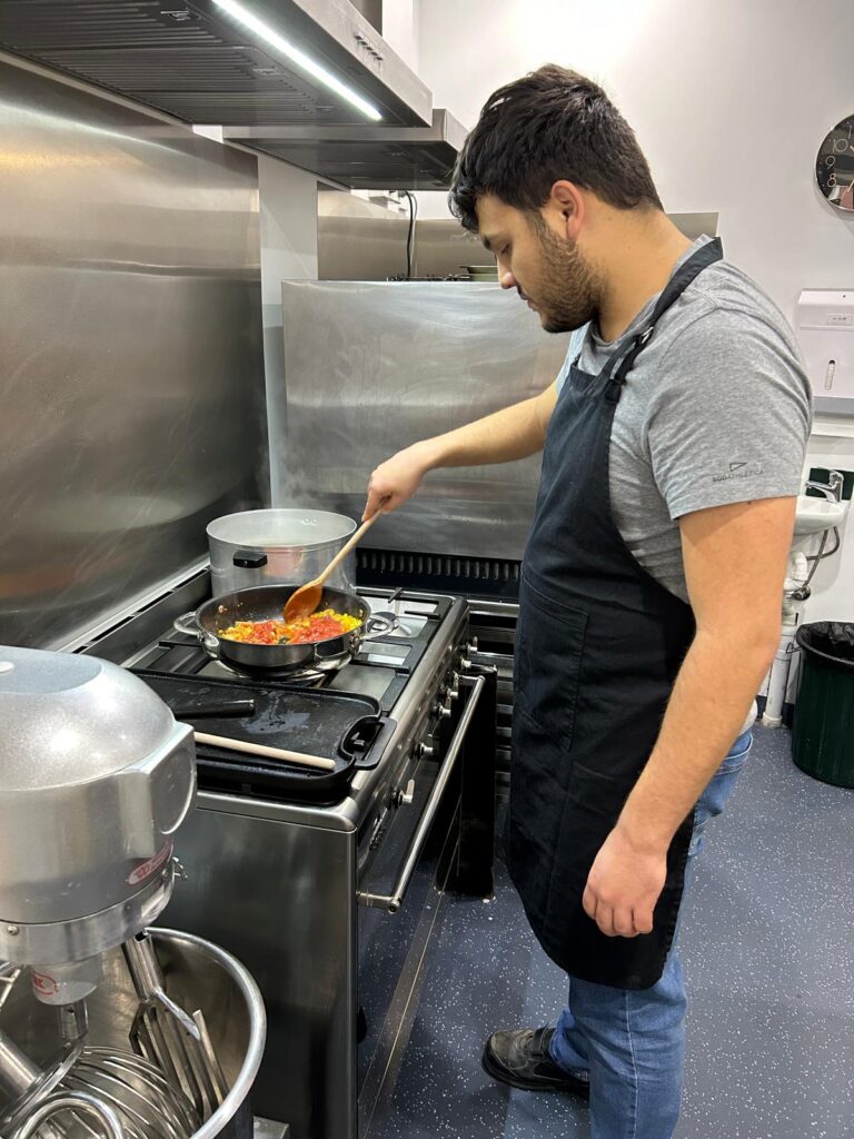 A Person Wearing A Black Apron And Gray T-Shirt Is Celebrating Culinary Success As They Cook At A Stove In A Modern Kitchen. They Are Stirring A Skillet Filled With Colorful Vegetables. The Kitchen Boasts Stainless Steel Appliances, Including An Industrial Mixer And A Large Pot On An Adjacent Burner.