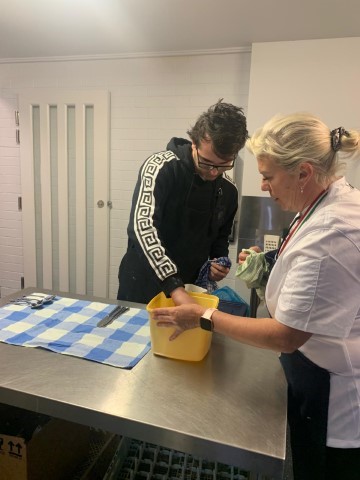 A Young Man And A Woman Are Standing In A Kitchen, Celebrating Their Culinary Success. The Man, Wearing A Black Hoodie With White Patterns On The Sleeves, Reaches Into A Yellow Container. The Woman, Dressed In A White Shirt And Holding A Cloth, Assists Him. A Blue Checkered Cloth Lies On The Counter.