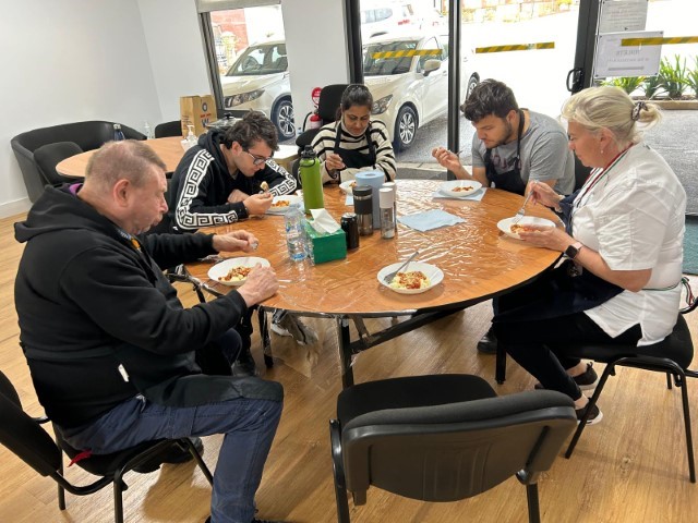 Five People Sit Around A Round Table, Celebrating With A Meal Together. The Table Is Covered With A Protective Sheet, And Plates Of Culinary Delights Are In Front Of Each Person. The Room Has Large Windows And A Wooden Floor, With A Couple Of Cars Visible Through The Windows Outside.