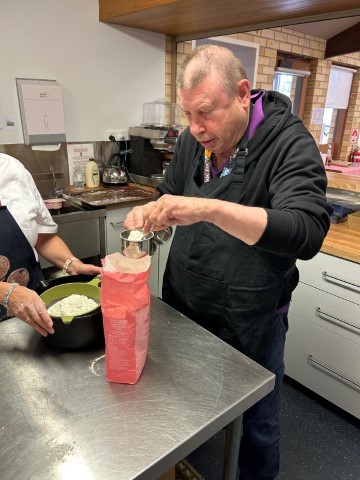 A Man Wearing A Black Apron Is Scooping Flour From A Bag Using A Measuring Cup In A Kitchen. Another Person, Partially Out Of Frame, Stands Beside Him Holding A Bowl Of Flour. Kitchen Appliances And Ingredients Are Visible In The Background, Creating The Perfect Setting For Their Culinary Success.