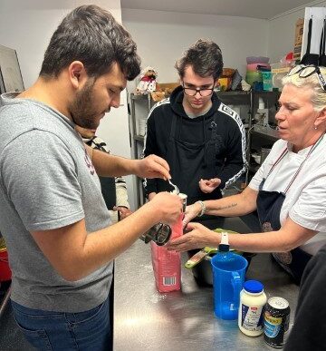 A Group Of People In A Kitchen Is Working Together On A Cooking Activity. One Person Is Using A Grater, Held Over A Pink Container, While Two Others Watch Closely. The Scene Of Culinary Success Unfolds In An Organized Kitchen With Various Utensils And Ingredients On The Counter.