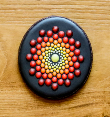 A Round, Decorated Cookie With A Black Background, Featuring A Pattern Of Small, Colored Dots Arranged In Concentric Circles. The Dots Transition From Yellow In The Center To Orange And Then To Red On The Outer Circles. It Is Placed On A Wooden Surface As Part Of Our Day Options Arts &Amp; Crafts Program For Individuals With Disabilities.