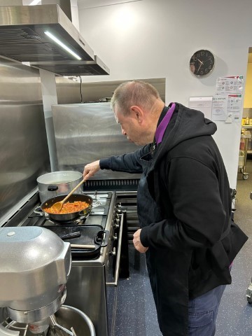 A Man Cooks In A Modern Kitchen. He Is Stirring Food In A Skillet On The Stove. The Kitchen Has Stainless Steel Appliances, A Pot On The Back Burner, And A Wall Clock In The Background. The Man Is Wearing A Black Hoodie And Apron.