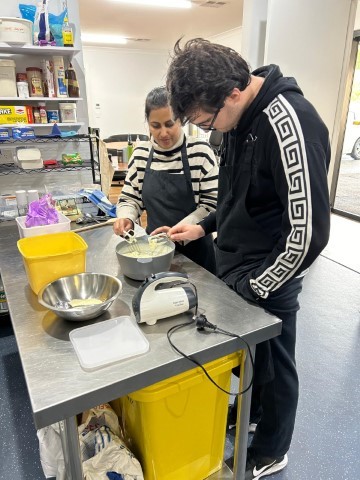 Two People Are In A Kitchen, Preparing Food. The Woman On The Left, Wearing A Black And White Striped Shirt, Is Stirring A Bowl. The Man On The Right, Wearing Black Pants And A Black Hoodie With A White Geometric Design On The Sleeves, Is Standing Next To Her. They Are Celebrating Their Culinary Success Together.