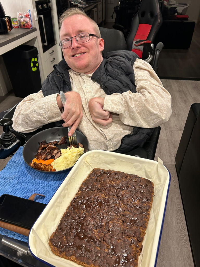 A Man In A White Shirt And Black Vest Sits In A Wheelchair At A Table. He Is Smiling, Holding A Fork, Enjoying Classic Dishes Like Meat, Mashed Potatoes, And Carrots. In Front Of Him Is A Large Tray Of Baked Dessert. This Scene Perfectly Captures The Essence Of Our Week 4 Update.