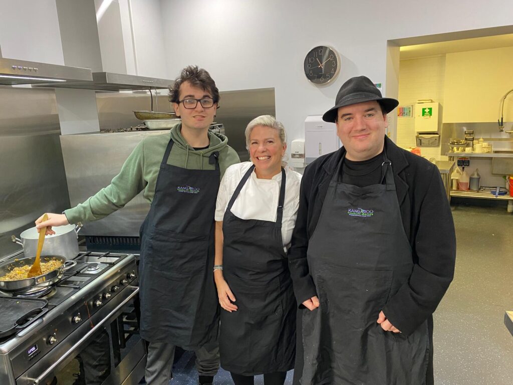 Three People Are Standing In A Kitchen, All Wearing Aprons. The Person On The Left Is Stirring Food In A Pan On The Stove. The Person In The Middle Is Smiling Warmly At The Camera, Showcasing Their Recipe For Success, While The Person On The Right Also Smiles. A Clock Is Visible In The Background.