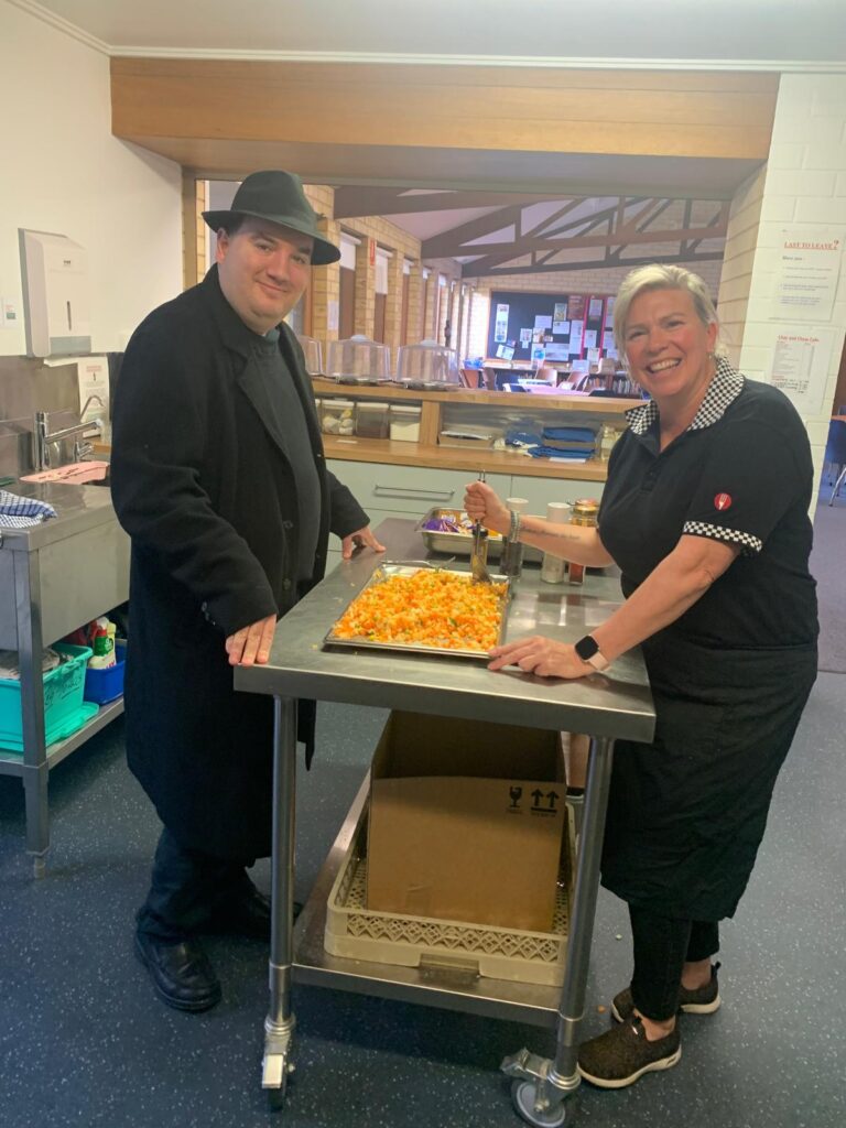 Two People Smiling While Standing On Either Side Of A Metal Table With A Tray Of Food. The Person On The Left Is Wearing A Black Coat And Hat, While The Person On The Right Is Dressed In A Black Uniform. They Appear To Be In A Kitchen Or Cafeteria Setting, Showcasing Their Culinary Highlights.