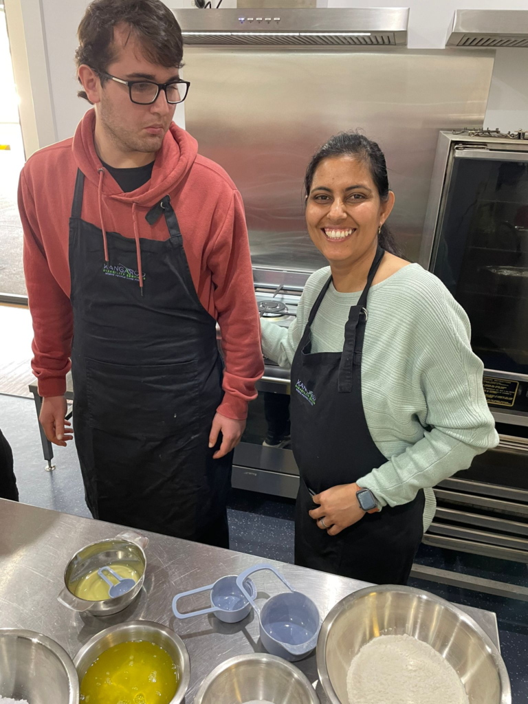 Two People In A Kitchen, Both Wearing Black Aprons, Stand By A Countertop With Several Metal Mixing Bowls Containing Ingredients. The Person On The Left Wears Glasses And A Red Hoodie, While The Person On The Right Smiles And Wears A Light Green Sweater. It'S Like They'Re Ready For Cooking Classes!