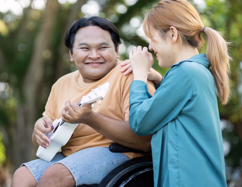 A Smiling Man In A Wheelchair Plays A White Ukulele While A Woman Stands Beside Him With Her Hand On His Shoulder. The Scene Is Set Outdoors, With Trees And Greenery Creating A Soft, Blurred Backdrop. The Man Wears A Gold-Colored Shirt And Denim Shorts, Showcasing A Casual Style. The Woman Is Dressed In A Long Teal Jacket. The Overall Composition Suggests A Light, Carefree Mood. This Image Would Be Well-Suited For Projects Related To Music, Companionship, Outdoor Activities, Or Accessible Lifestyles.