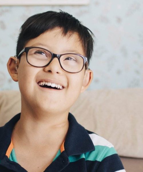 A Young Boy With Short Dark Hair And Glasses Is Smiling While Sitting On A Couch. He Is Wearing A Striped Green And Blue Shirt. The Background Features Light-Colored, Patterned Wallpaper, Emphasizing A Warm And Welcoming Space Perfect For An Ndis Provider Dedicated To Supporting Those With Disabilities.
