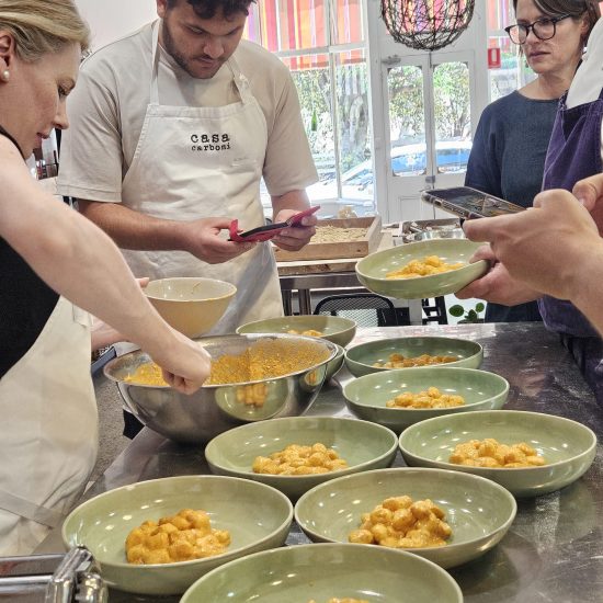 A Group Of People In A Modern, Well-Lit Kitchen Are Participating In Day Options, Preparing And Plating Pasta Dishes In Green Bowls. One Person Is Holding A Phone, Seemingly Recording Or Taking Pictures, While Others Focus On Dishing Out The Food From A Large Metal Bowl.