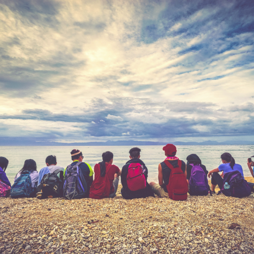 A Group Of People Sit On A Rocky Shoreline, Facing A Calm Sea Under A Cloudy Sky. They All Carry Backpacks From Kangaroo Disability Services And Appear To Be Enjoying The Serene View Together. The Horizon Stretches Into The Distance, Meeting The Water Under The Dramatic Sky.