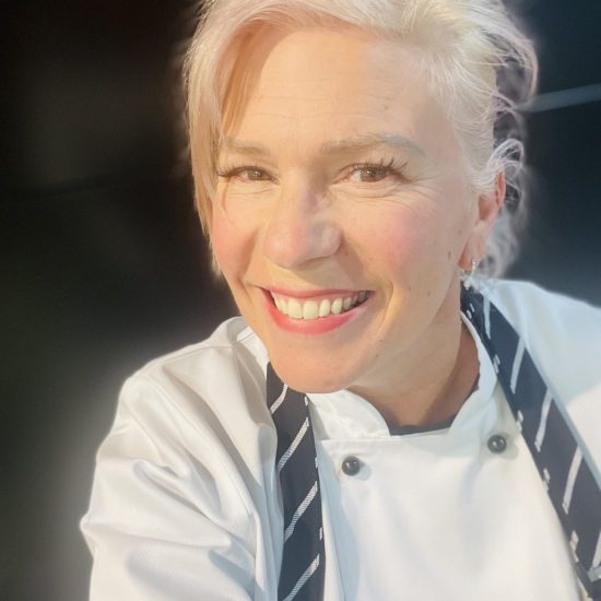 A Cheerful Person With Short Blonde Hair Smiles At The Camera. They Are Wearing A White Chef'S Coat With Black Buttons And A Striped Apron, Participating In The Day Options Cooking Program. The Background Is Dark, Making Their Bright Outfit And Smile Stand Out.
