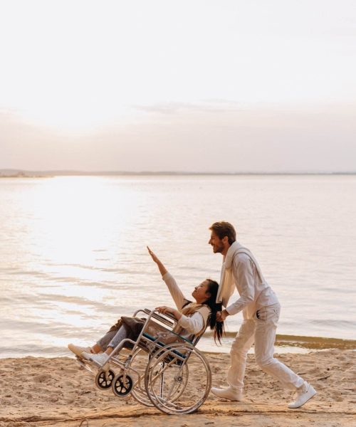 A Person, Possibly An Ndis Provider, Is Pushing Another In A Wheelchair Along A Sandy Beach At Sunset. The Person In The Wheelchair Is Smiling And Reaching Out Towards The Sky, Capturing A Joyful And Carefree Moment By The Water Despite Their Disability.