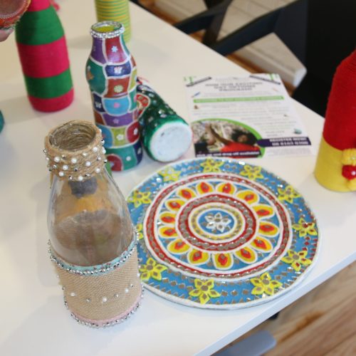 A Table Displaying Various Decorated Items, Including Colorful Handmade Bottles Wrapped In Yarn, Embellished With Beads And Fabric, And A Decorative Plate With Intricate Patterns And Designs. Brochures Detailing May Events Are Also Visible In The Background.