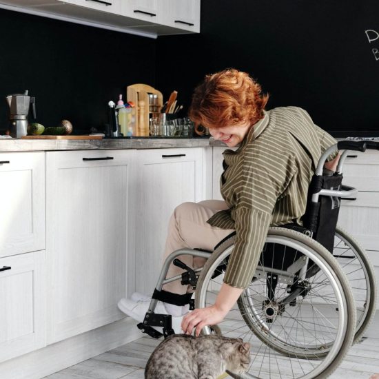 A Person With A Disability, Supported By Their Ndis Provider, Is In A Kitchen With White Cabinets And A Black Wall. They Are Reaching Down To Pet A Gray Cat That Is Standing On The Floor. The Person Has Short, Curly Hair And Is Wearing A Striped Shirt. Various Kitchen Items Are Visible On The Counters.