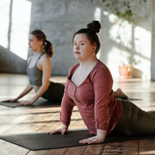 Two Women Practice Yoga Indoors On Mats In A Well-Lit Room With Wooden Floors, Demonstrating The Upward-Facing Dog Pose. One Woman In The Foreground Wears A Red Top, The Other In Grey, With Sunlight Streaming Through Large Windows—A Serene Environment Perfect For All Abilities.