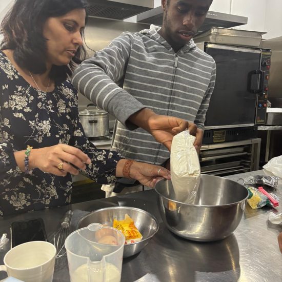 A Woman And A Man Are Cooking In A Kitchen. The Man Pours Ingredients From A Packet Into A Metal Bowl. Various Cooking Ingredients And Utensils Are On The Table. Both Are Focused On Their Activity. A Woman And A Man Are Cooking In A Kitchen. The Man Pours Ingredients From A Packet Into A Metal Bowl. Various Cooking Ingredients And Utensils Are On The Table. Both Are Focused On Their Activity.