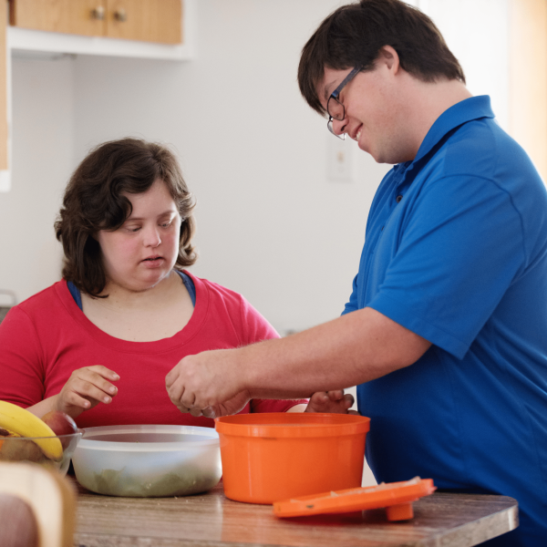 Two People With Down Syndrome Are In A Kitchen, Joyfully Preparing Food Together As Part Of Their Day Options. The Person On The Right In A Blue Shirt Stirs Contents In An Orange Container, While The Person On The Left In A Red Shirt Looks On. There Are Fruits And A Bowl On The Counter In The Foreground.