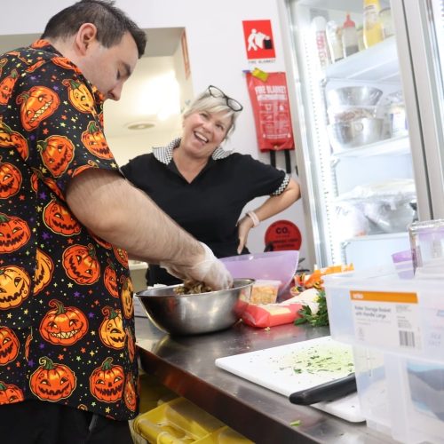A Person In A Halloween-Themed Shirt Prepares Mexican Food, Mixing Ingredients In A Bowl. Another Person Standing Nearby Smiles While Watching, Much Like A Scene From A Cooking Program. The Background Shows A Refrigerator And Various Kitchen Items. A Person In A Halloween-Themed Shirt Prepares Mexican Food, Mixing Ingredients In A Bowl. Another Person Standing Nearby Smiles While Watching, Much Like A Scene From A Cooking Program. The Background Shows A Refrigerator And Various Kitchen Items.