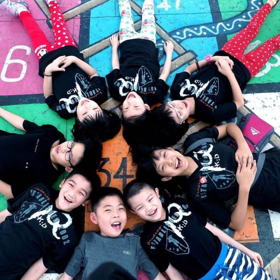 A Group Of Eight Children, Dressed In Black T-Shirts And Colorful Pants, Lie On A Hopscotch Court In A Circle With Their Heads Touching In The Center. They Are Smiling And Laughing, Creating A Playful And Joyful Atmosphere.