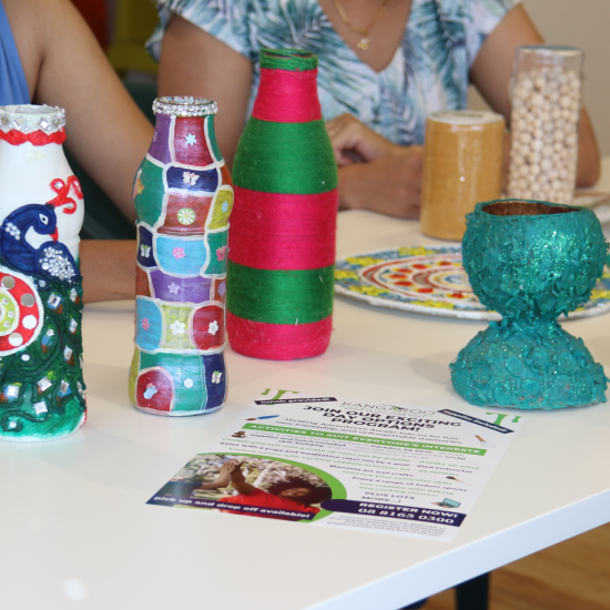 Decorated Bottles Are Displayed On A Table, Each Featuring Unique Designs, Colors, And Textures Like Sequins And Paint. Part Of The Day Options Arts &Amp; Crafts Program For Individuals With Disabilities, There Are Craft Flyers On The Table, And Two People Are Partially Visible In The Background.