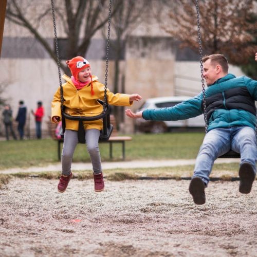 A Child And An Adult Are Sitting On Swings In A Park, Embodying The Spirit Of Our Mission. The Child, In A Red Hat And Yellow Jacket, Reaches Out To The Teal-Clad Adult. They Smile, Clearly Enjoying Their Time Together. Trees And People Fill The Background, Illustrating The Community We Cherish.