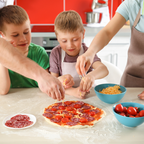 Two Children And Two Adults, All Wearing Aprons, Are Preparing A Homemade Pizza In A Kitchen As Part Of The Day Options Kids Cooking Program. The Children Are Adding Cheese And Toppings While The Adults Assist. A Plate Of Sliced Salami And A Bowl Of Cherry Tomatoes Are Nearby On The Floured Countertop.