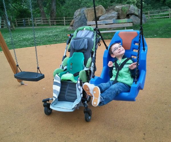 A Child Wearing Glasses And A Green Jacket Is Joyfully Swinging In A Blue Adaptive Swing Next To An Empty Regular Swing. A Stroller With A Green Seat Is Positioned Nearby On The Playground With A Grassy Area And Rocks In The Background.