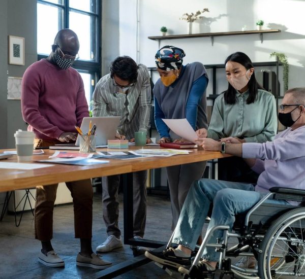 A Diverse Group Of Five People, All Wearing Masks, Collaborate Around A Table In A Modern Office. One Person Uses A Wheelchair, While The Others Are Standing And Looking At Documents And A Laptop. Various Papers Related To Ndis And A Coffee Cup Are Spread Out On The Table.