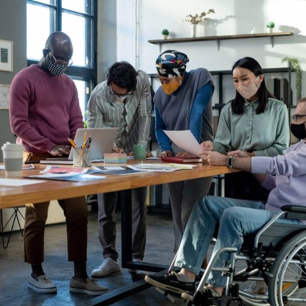A Diverse Group Of Five People, All Wearing Masks, Collaborate Around A Table In A Modern Office. One Person Uses A Wheelchair, While The Others Are Standing And Looking At Documents And A Laptop. Various Papers Related To Ndis And A Coffee Cup Are Spread Out On The Table.